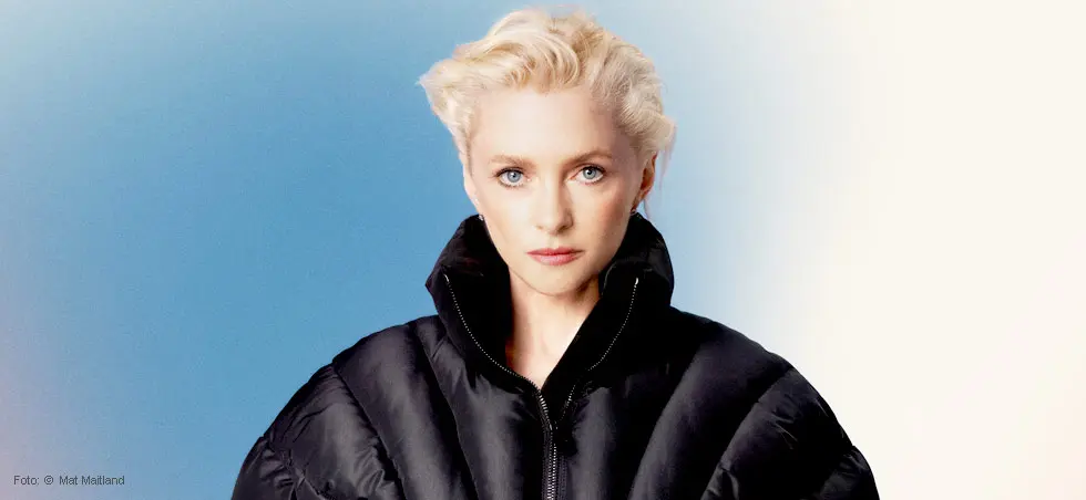 Alison Goldfrapp mit blondem Haar und schwarzer Jacke vor blau-weißem Hintergrund.