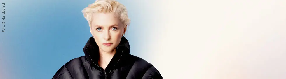 Alison Goldfrapp mit entschlossenem Blick, trägt eine schwarze Jacke. Hintergrund ist blau-weiß verschwommen.