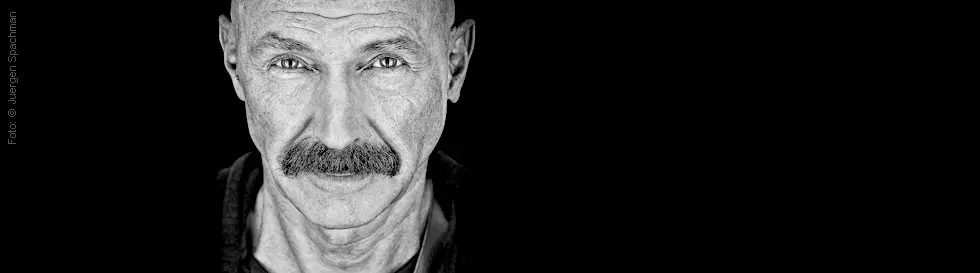 Schwarz-weiß Porträt von Tony Levin mit Glatze und Schnurrbart, direkt in die Kamera blickend.