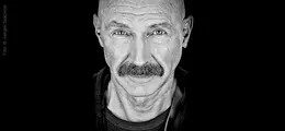 Schwarz-weiß Porträt von Tony Levin mit Glatze und Schnurrbart, direkt in die Kamera blickend.