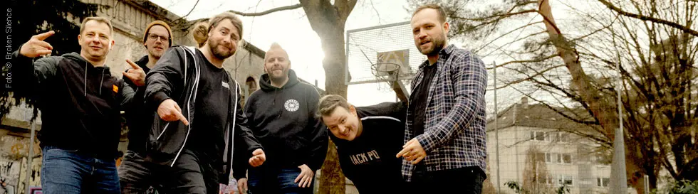 Sechs Männer posieren lässig auf einem Basketballfeld mit Graffiti im Hintergrund.