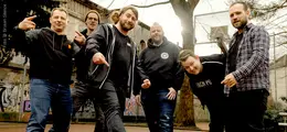 Sechs Männer posieren lässig auf einem Basketballfeld mit Graffiti im Hintergrund.