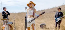 Orianthi Panagaris mit Gitarre steht draußen auf einem Feld, flankiert von einem Schlagzeuger und einem Bassisten.