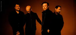 Eine Gruppe von vier Männern steht vor einem orange-braunen Hintergrund. Alle tragen dunkle Kleidung.
