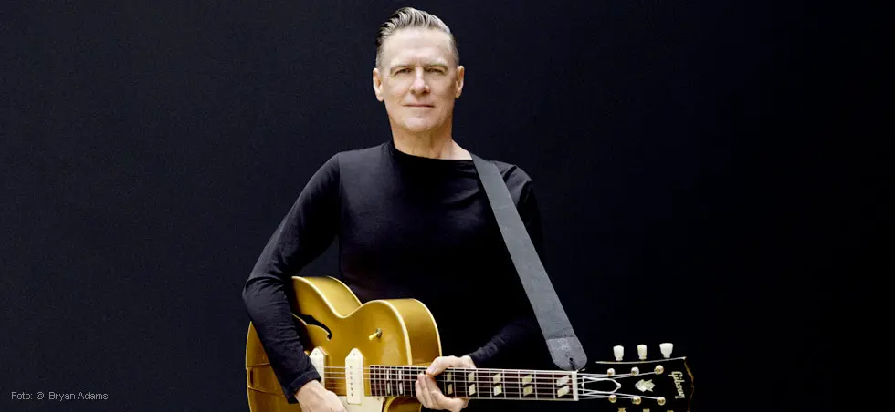 Eine Person in schwarz hält eine goldene Gitarre vor einem schwarzen Hintergrund.