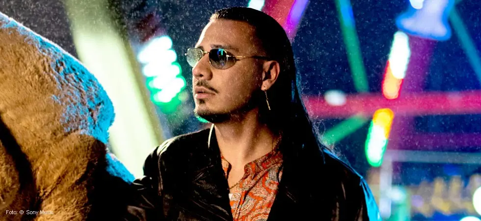Ein Mann mit Sonnenbrille in lebhafter Umgebung, bunte Lichter im Hintergrund.