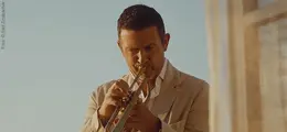 Ein Mann in einem beigen Anzug spielt eine Trompete vor einem Fenster bei warmem, goldenen Licht.