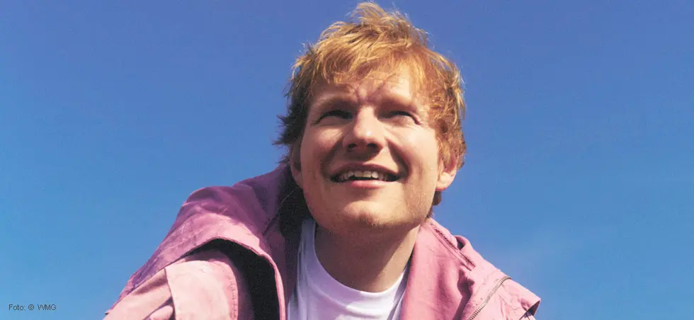 Ein lächelnder Mann mit rötlichen Haaren trägt eine rosa Jacke vor einem blauen Himmel.