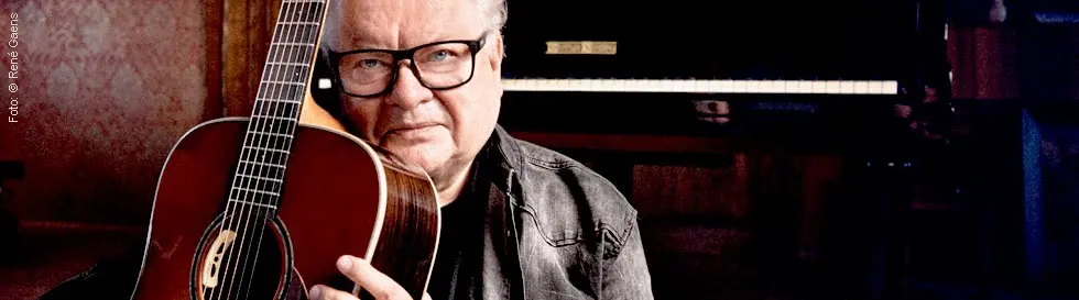 Ein älterer Mann mit Brille hält eine Akustikgitarre. Im Hintergrund ist ein Klavier sichtbar.