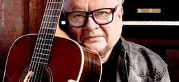 Ein älterer Mann mit Brille hält eine Akustikgitarre. Im Hintergrund ist ein Klavier sichtbar.