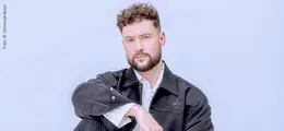 Ein Mann mit lockigem Haar und Bart schaut ernst. Er trägt eine dunkle Jacke und sitzt vor hellem Hintergrund.