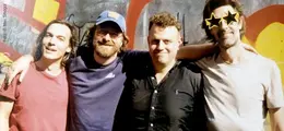 Vier Männer lachen in Freizeitkleidung vor bunter Wand. Einer trägt eine sternförmige Sonnenbrille.