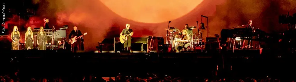 Vier Musiker auf einer Bühne, einer spielt Gitarre, ein anderer sitzt am Schlagzeug, warme Beleuchtung.