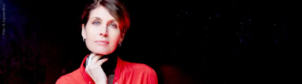 Frau in roter Jacke, Hand am Kinn, ruhiger Ausdruck, dunkler Hintergrund.
