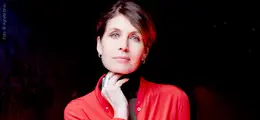 Frau in roter Jacke, Hand am Kinn, ruhiger Ausdruck, dunkler Hintergrund.