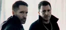 Zwei Männer in dunkler Kleidung blicken ernst in die Kamera. Die Szene ist in weichem Licht gehalten.