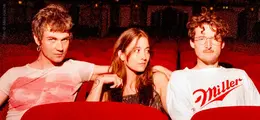 Zu sehen sind drei Personen in einem Theater. Rechts trägt eine Person ein weißes T-Shirt.