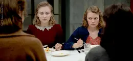 Zwei Frauen sitzen an einem Tisch, essen und wirken angespannt. Sie tragen elegante Kleidung und blicken ernst.