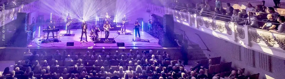 Eine Band auf einer Bühne mit farbigen Lichtern spielt vor einem sitzenden Publikum in einem Konzertsaal.