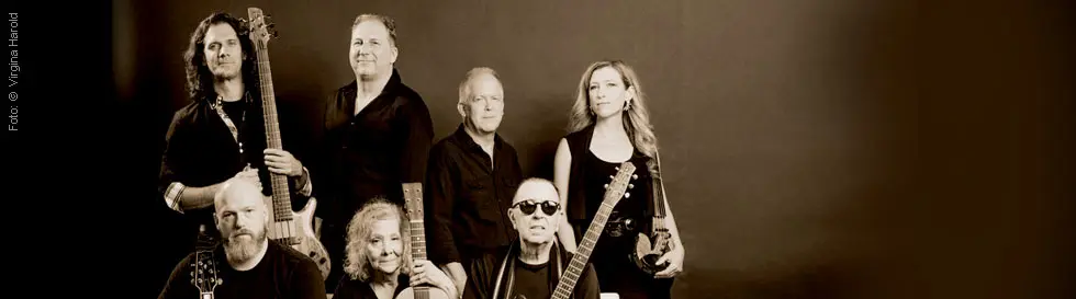 Sieben Personen mit Musikinstrumenten posieren zusammen, lächeln oder schauen ernst. Sepia-Ton.