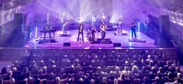 Eine Band auf einer Bühne mit farbigen Lichtern spielt vor einem sitzenden Publikum in einem Konzertsaal.
