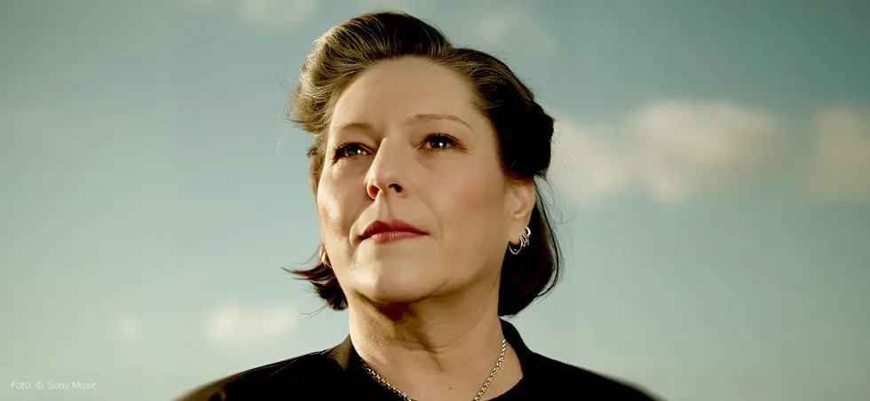 Eine Person blickt in die Ferne, mit kurzem dunklem Haar und Ohrringen, vor einem Himmel mit wenigen Wolken.