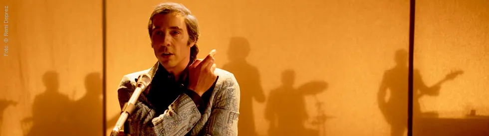 Ein Mann im gemusterten Anzug singt ins Mikrofon, Schatten von Musikern im Hintergrund.