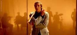 Ein Mann im gemusterten Anzug singt ins Mikrofon, Schatten von Musikern im Hintergrund.