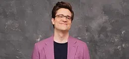Ein Mann mit Brille und lila Sakko vor grauem Hintergrund.