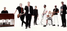 Mehrere Männer in formeller und Freizeitkleidung, mit Musikinstrumenten vor weißem Hintergrund.