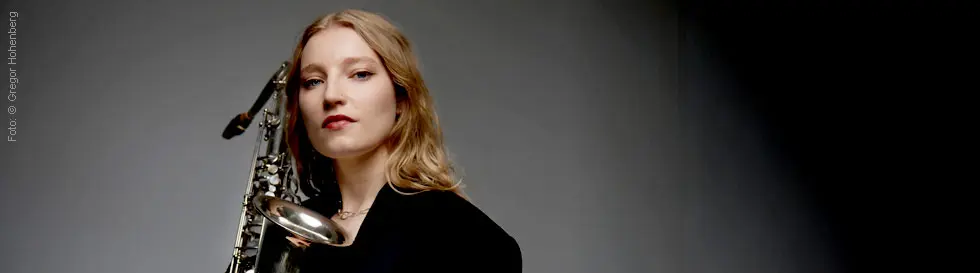 Eine Frau mit blondem Haar hält ein Saxofon, sie trägt schwarze Kleidung. Der Hintergrund ist grau.