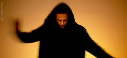 Eine Person mit Kapuze, Kopf gesenkt, in einer unscharfen Bewegung vor orangefarbenem Hintergrund.