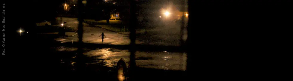 Ein nächtliches Szenario zeigt eine einsame Gestalt in der Ferne auf einer nassen Straße unter Straßenlaternenlicht.