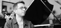 Eine Person mit Brille sitzt vor einem Flügel, im Hintergrund sind unscharfe Elemente zu erkennen.