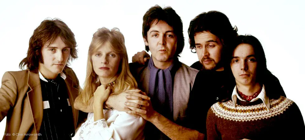 Fünf Menschen stehen nebeneinander vor einem weißen Hintergrund. Sie blicken direkt in die Kamera.