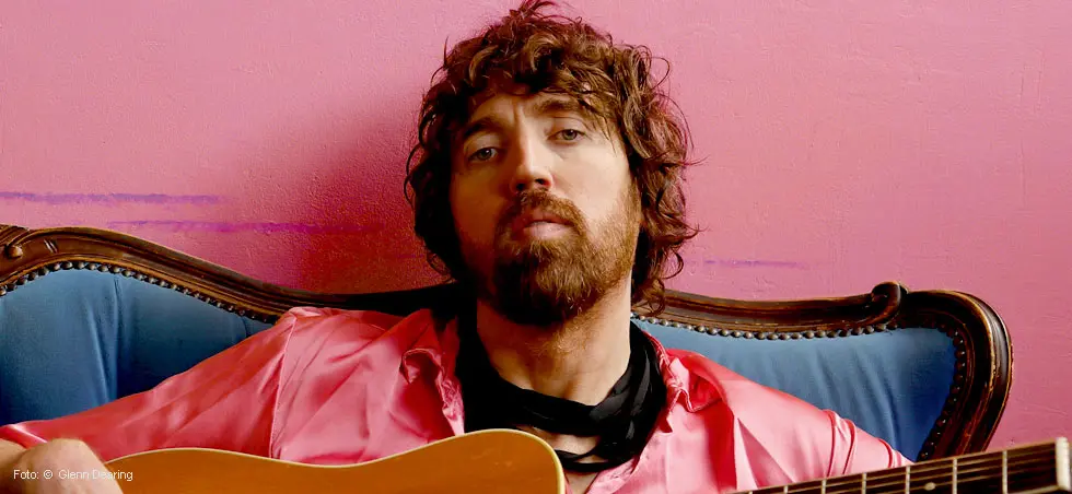 Eine Person mit Bart sitzt auf einem blauen Sofa vor einer rosa Wand, trägt ein rosa Hemd und hält eine Gitarre.