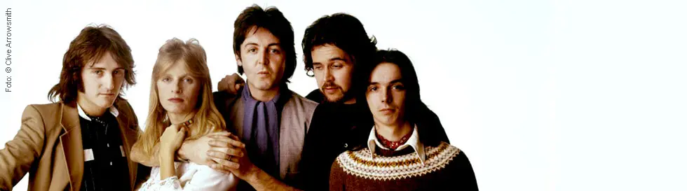 Eine Gruppe von fünf Personen, eng nebeneinander stehend. Unterschiedliche Kleidung; entspannt und freundlich.