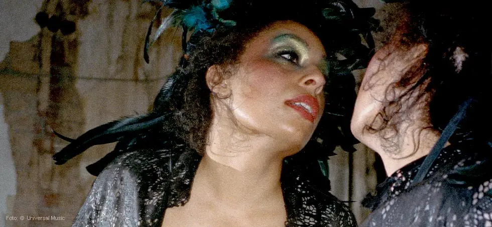 Eine Frau mit leuchtendem Make-up und Federschmuck schaut in einen Spiegel.