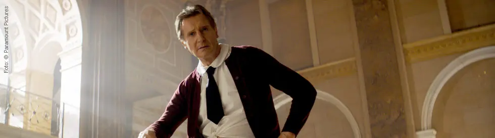 Eine Person steht in einem eleganten Raum, trägt Hemd und Krawatte, aber keine Hosen sondern einen karierten Rock und eine gepunktete Unterhose.