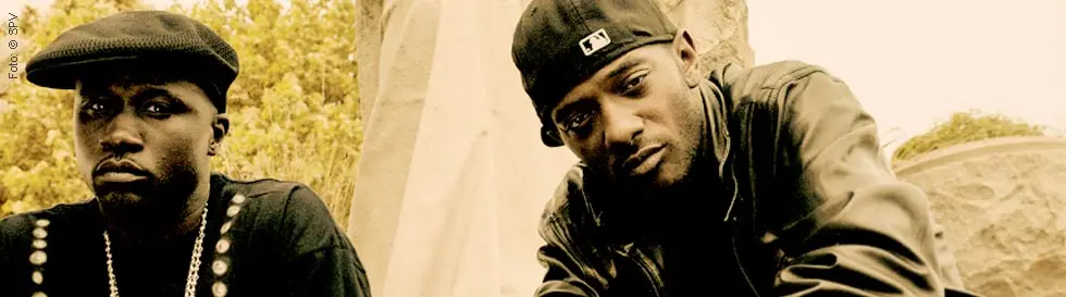 Zwei Männer in dunkler Kleidung und Ketten, vor einer Statue posierend.