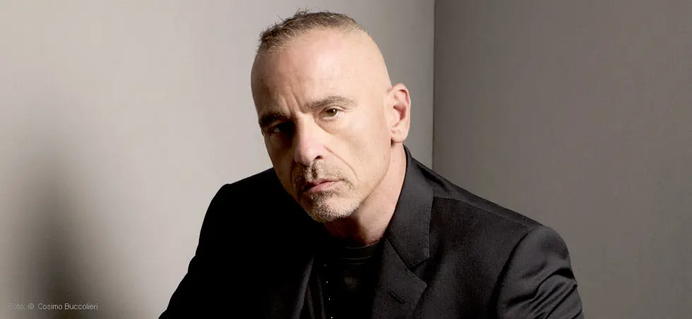 Eine nachdenklich dreinblickende Person in dunkler Kleidung vor einem neutralen Hintergrund.