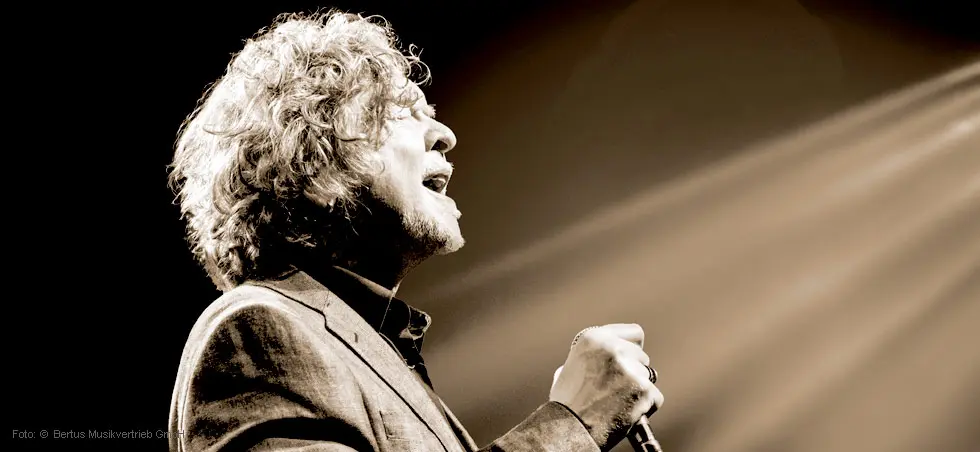 Ein Mann mit lockigem Haar singt leidenschaftlich in ein Mikrofon, angeleuchtet von einem hellen Scheinwerfer.