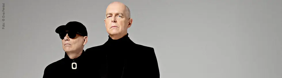 Zwei Männer in Schwarz, einer trägt eine Mütze und Sonnenbrille, der andere hat einen ernsten Ausdruck und ist glatzköpfig.
