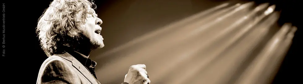 Ein Sänger mit lockigem Haar singt leidenschaftlich in ein Mikrofon, dramatisch beleuchtet.