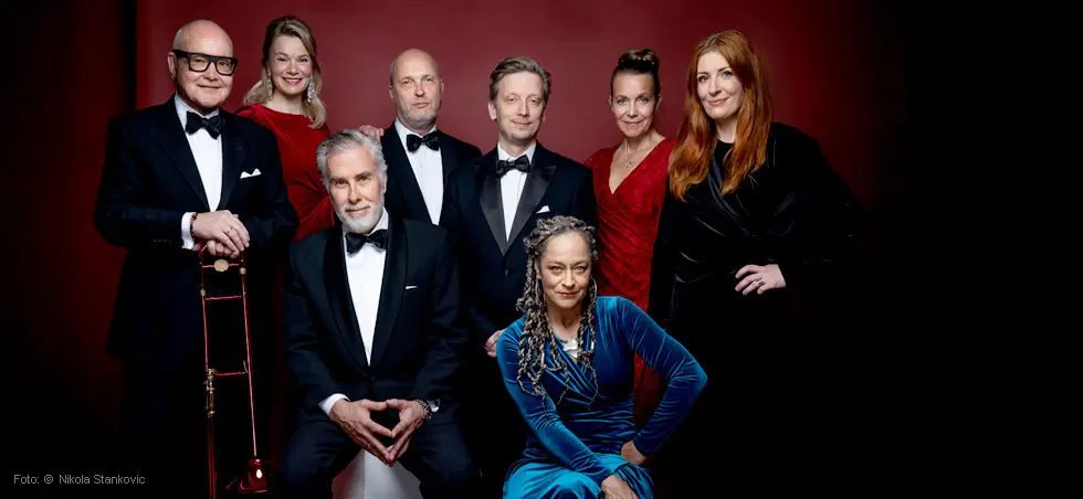 Acht elegant gekleidete Personen in Abendgarderobe posieren vor einem tiefroten Hintergrund.