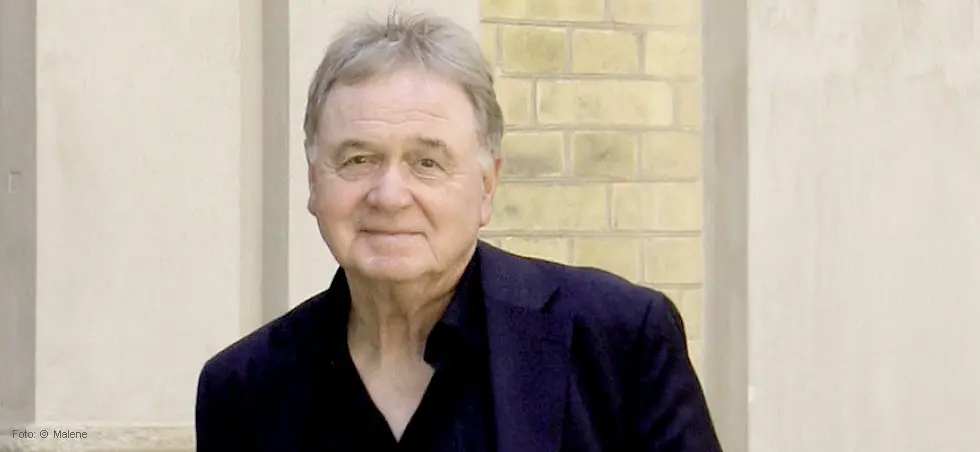 Ein älterer Mann in dunklem Sakko steht neben einer hellen, strukturierten Wand und blickt freundlich in die Kamera.