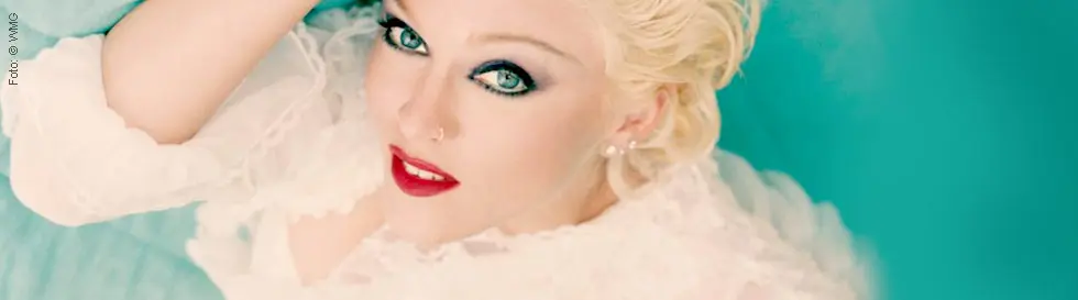 Die Person hat blondes Haar, roten Lippenstift und trägt ein weißes, geschichtetes Oberteil. Der Hintergrund ist türkis.