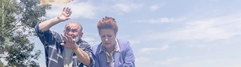 Ein älteres Paar im Freien, das nach fliegendem Geld greift. Links ein Baum, im Hintergrund hügelige Landschaft.