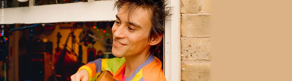 „Foto: Theo Batterham.“ Ein lächelnder Mensch mit einer bunten Jacke spielt Gitarre vor einem offenen Fenster.