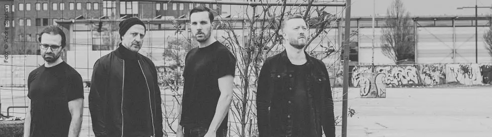 Vier Männer in schwarzer Kleidung stehen in einer urbanen, leicht heruntergekommenen Umgebung.